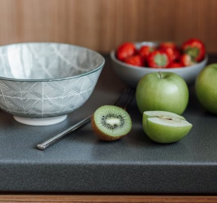 Table with bowl near fork and green apples with kiwi and cutting board near strawberries in dishware in light kitchen