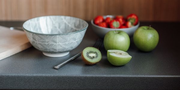 Table with bowl near fork and green apples with kiwi and cutting board near strawberries in dishware in light kitchen